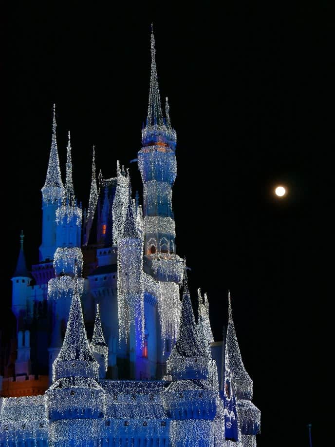 Cinderella's Castle at night lit up in white with the moon in the background at the Magic Kingdom in Disney World at Christmas