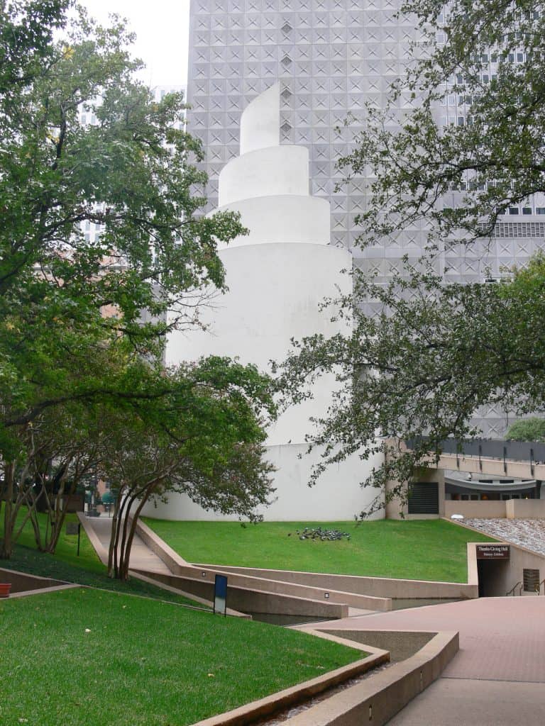 A white stone chapel that looks like an orange peel shape in the middle of green space with trees around