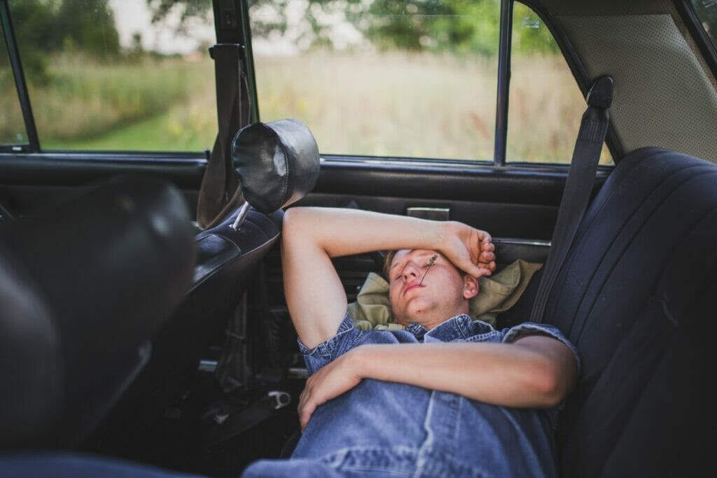 A person reclining in the back seat of a car, resting with their arm over their forehead.