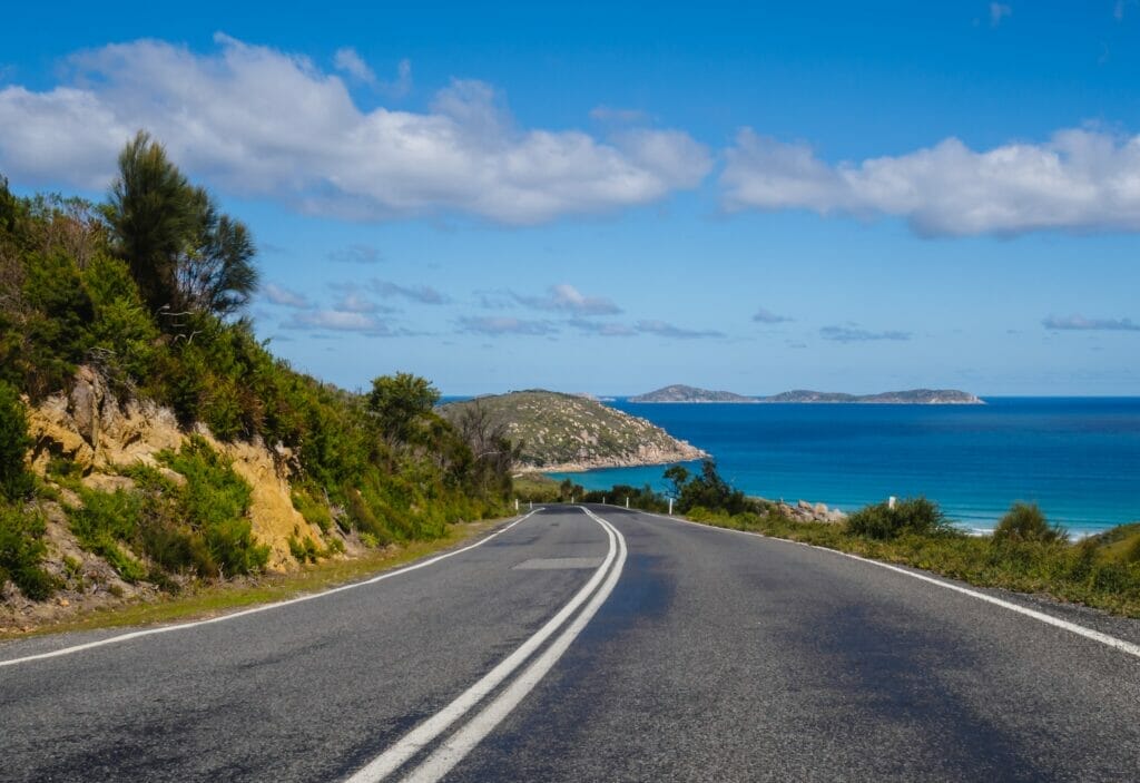 A two-lane highway along a beautiful coast, with blue waters to the right and green mountainside to the left.