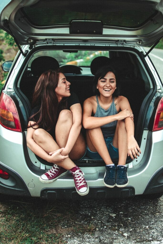 Two smiling people sitting on the rear bumper of a car with the back hatch open.