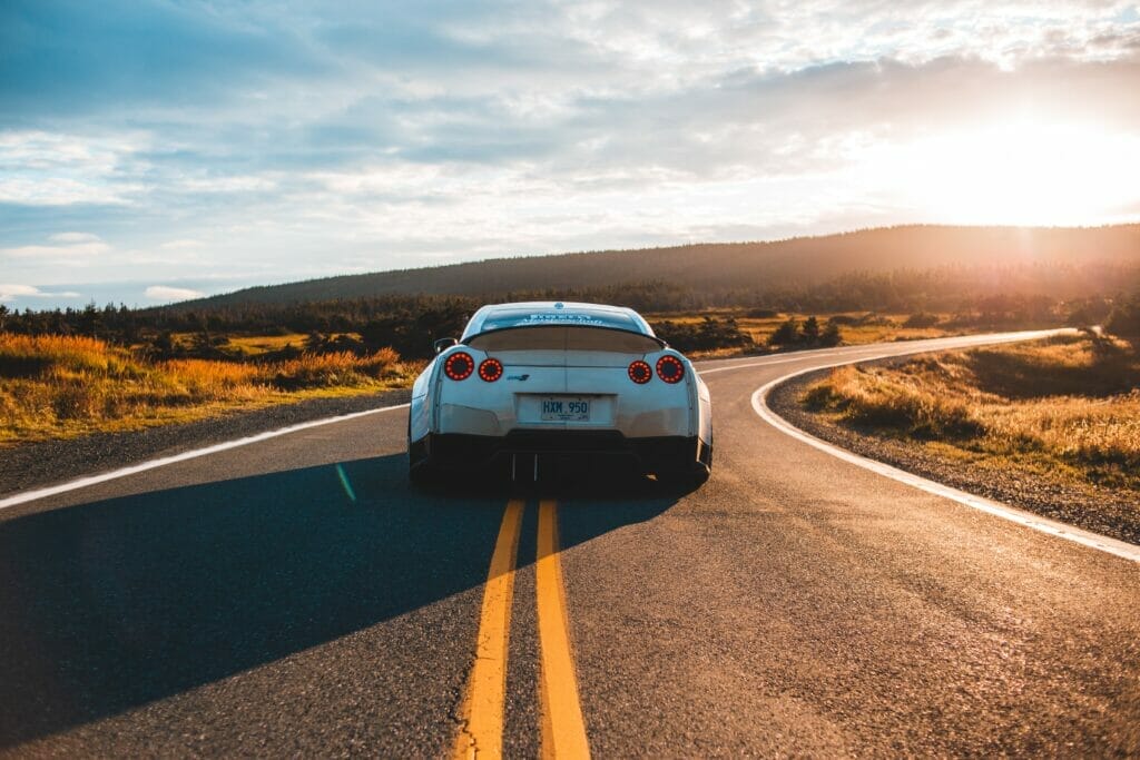 A car drives in the middle of a two lane highway, the bright sun in the distance behind a mountain range.