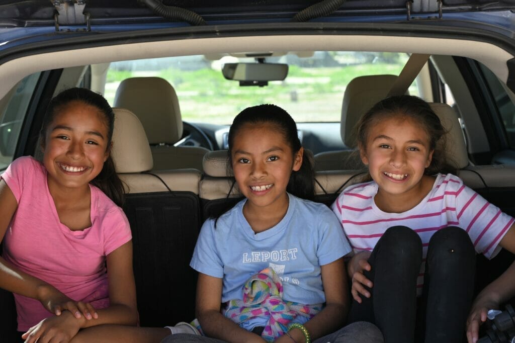 Three children sit in a row in the back of a car, smiling