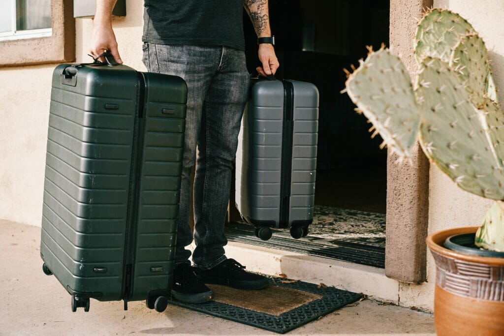A person holds two suitcases, standing on their front door step as if ready to leave for a flight