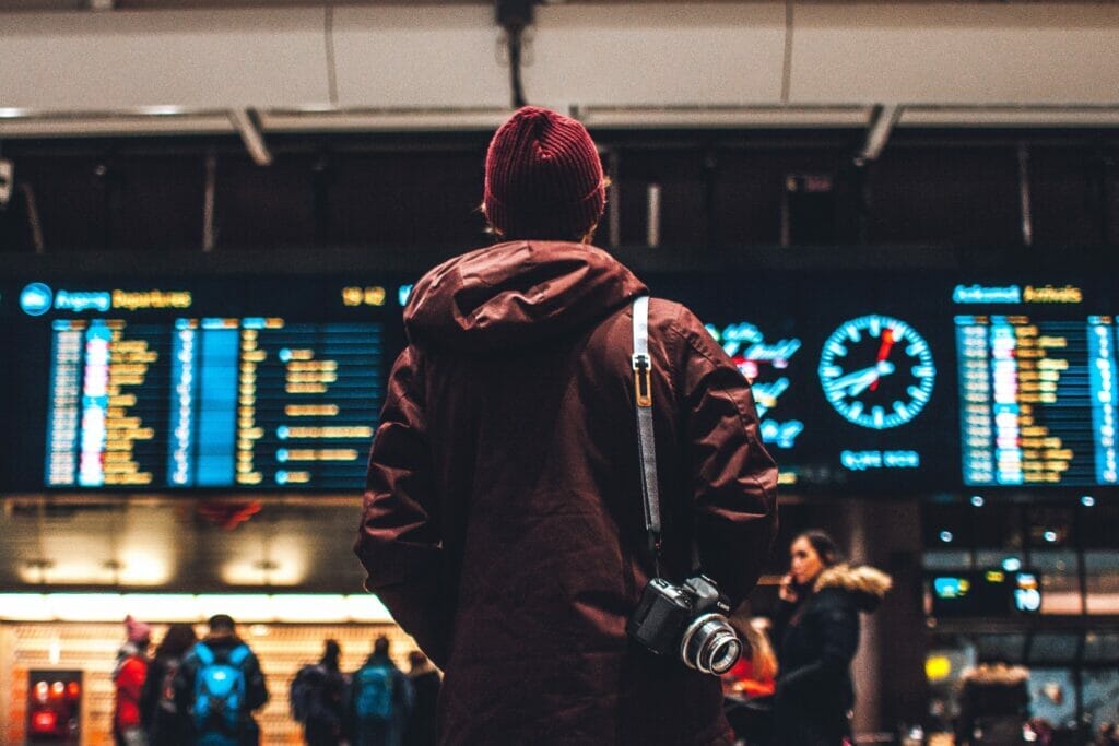 A person in winter clothing stands in an airport looking at the departure and arrival screens.