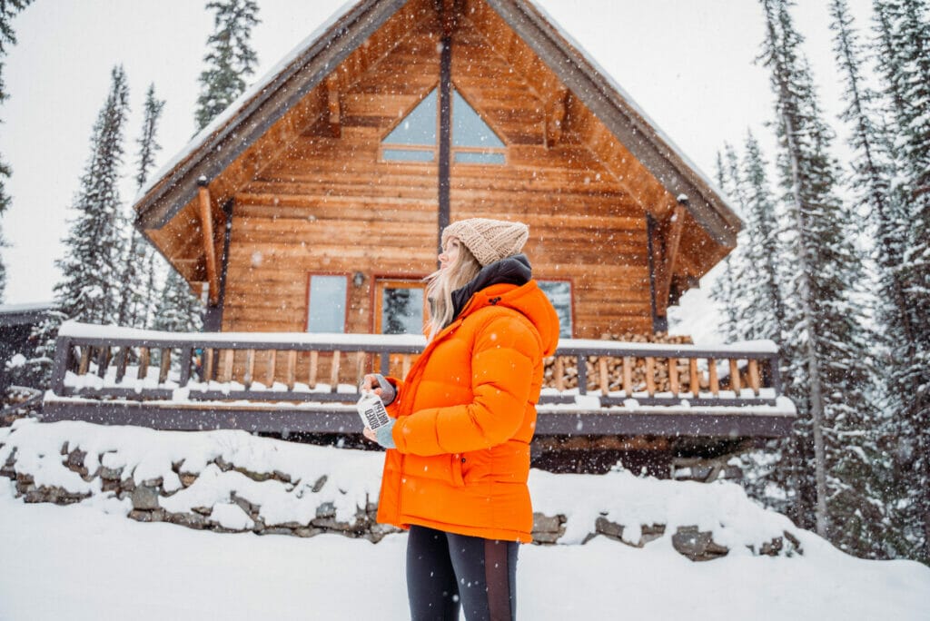 woman in orange jacket standing in front of a cabin in the snow