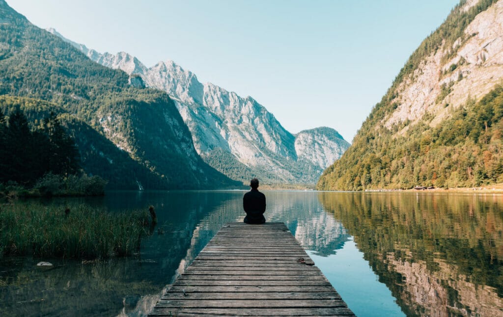 man sitting on the end of a dock looking at mountains.
