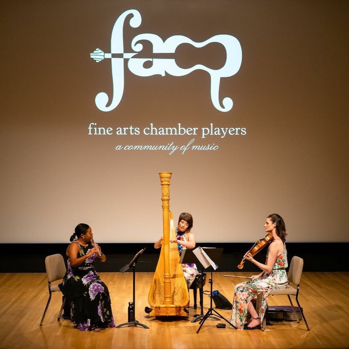 Three musicians on stage with the logo and title of the Fine Arts Chamber Players on a screen behind them