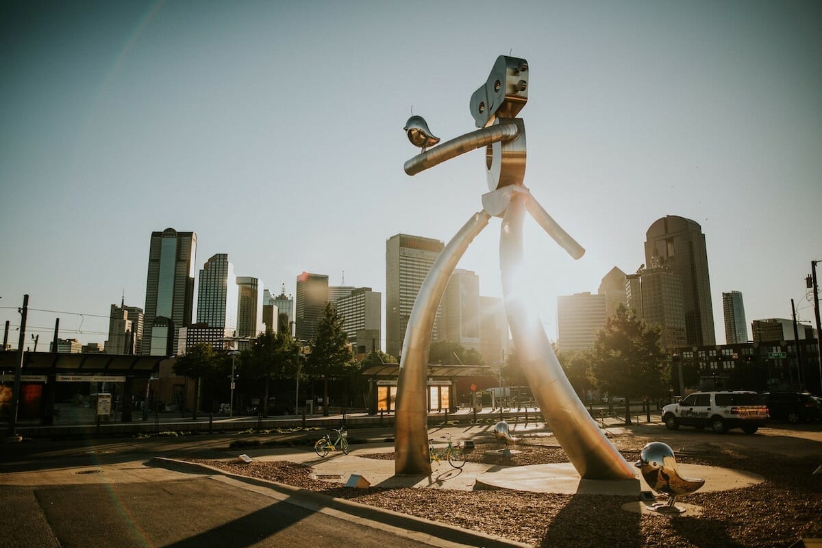 A tall metal sculpture of a robot-looking creature stands in a public square amidst the streets of Deep Ellum in Dallas Texas