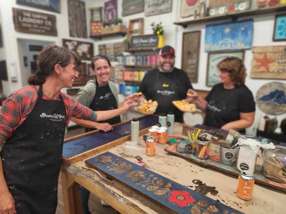 Four people smile and share snacks around a woodworking table at Board and Brush Duluth, MN